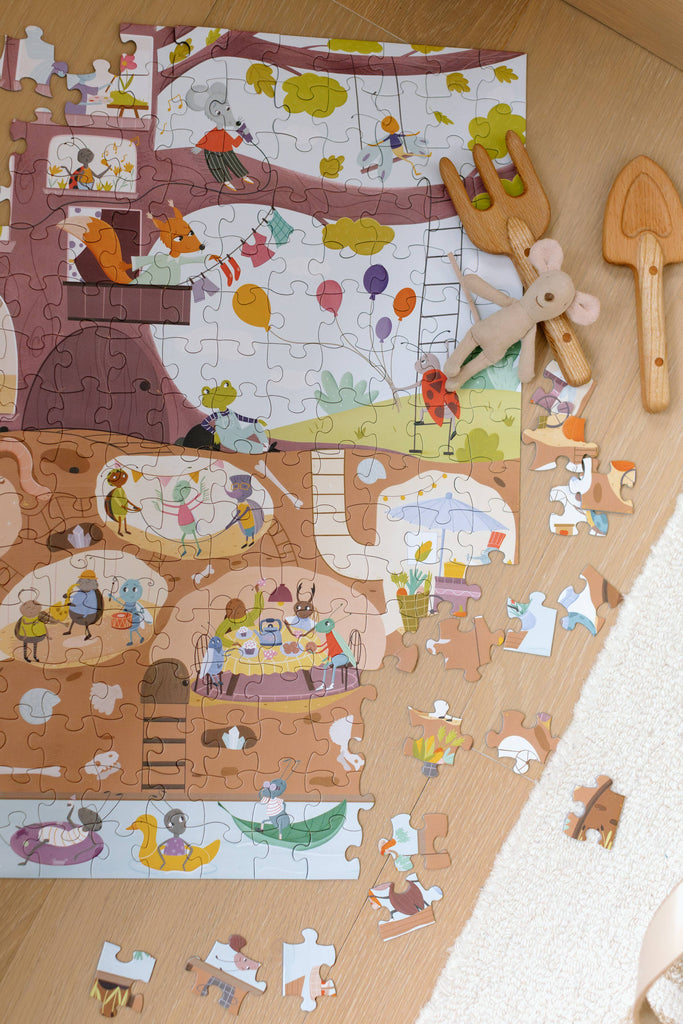 The Beneath the Branches 200 Piece Puzzle with animal characters lies partially assembled on wood beside a plush mouse and two wooden utensils. Matte pieces, some loose, are scattered near the edge of a white rug.