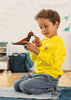 A young child in a yellow shirt kneels on a rug, holding and examining the Schleich Carnotaurus Large Realistic Dinosaur Toy With Movable Jaw with curiosity in a brightly lit room decorated with dinosaur wall decals, turning playtime into an engaging educational experience.