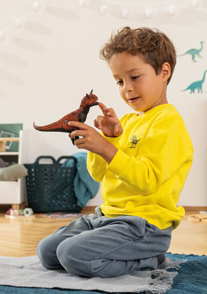 A young child in a yellow shirt kneels on a rug, holding and examining the Schleich Carnotaurus Large Realistic Dinosaur Toy With Movable Jaw with curiosity in a brightly lit room decorated with dinosaur wall decals, turning playtime into an engaging educational experience.