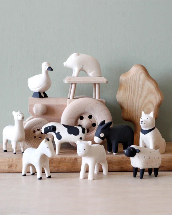 A collection of handcrafted wooden animal toys including a swan, bear, llama, fox, two sheep, and a donkey on a display with carved trees in the background.
