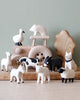 A collection of handmade wooden animal figurines, including a bear, birds, and various Handmade Tiny Wooden Farm Animals - Horse, displayed in front of a green wall, arranged artistically around a wooden structure.