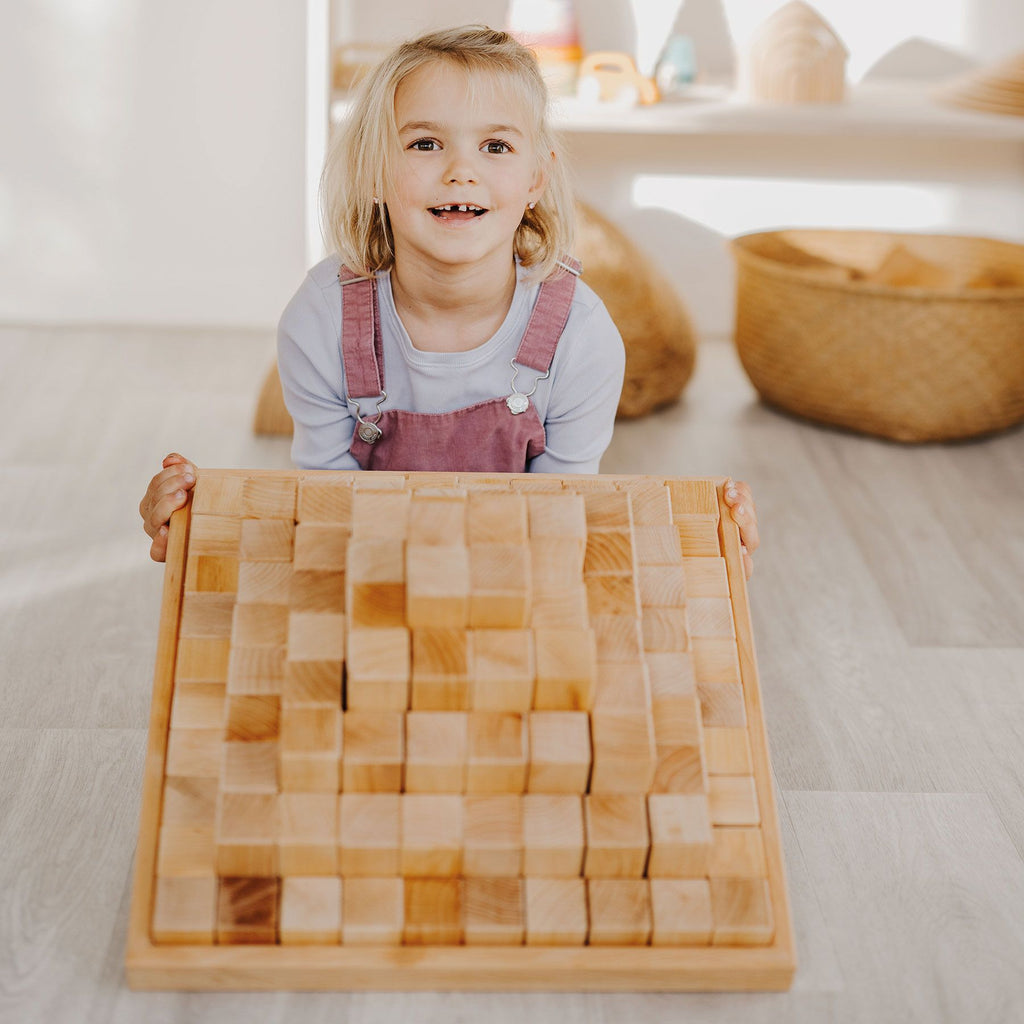 A young child with blond hair, wearing a light blue shirt and pink overalls, smiles while holding a wooden tray filled with Grimm's Large Natural Stepped Pyramid. The background shows a brightly lit room with woven baskets and shelves.