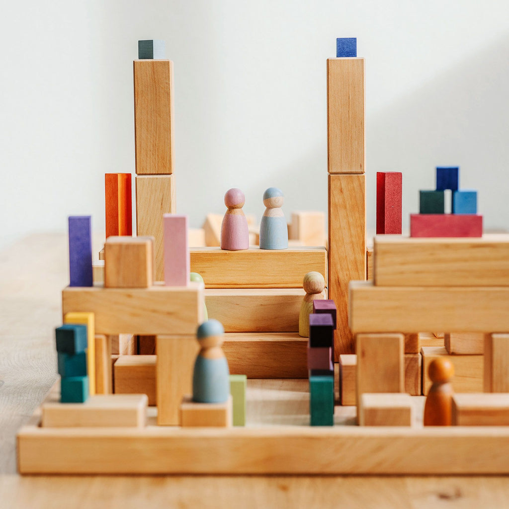 Grimm's Large Natural Stepped Pyramid, including a set of 100 building blocks, is arranged on a wooden surface to create small structures and platforms. Peg-shaped wooden figures in various colors are scattered around, forming what resembles a small abstract community.