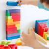 A child stacks Grimm's Stepped Counting Blocks - Small (coming soon) by color, with more wooden blocks and numbers on a white surface, creating a playful, non-toxic toy scene.