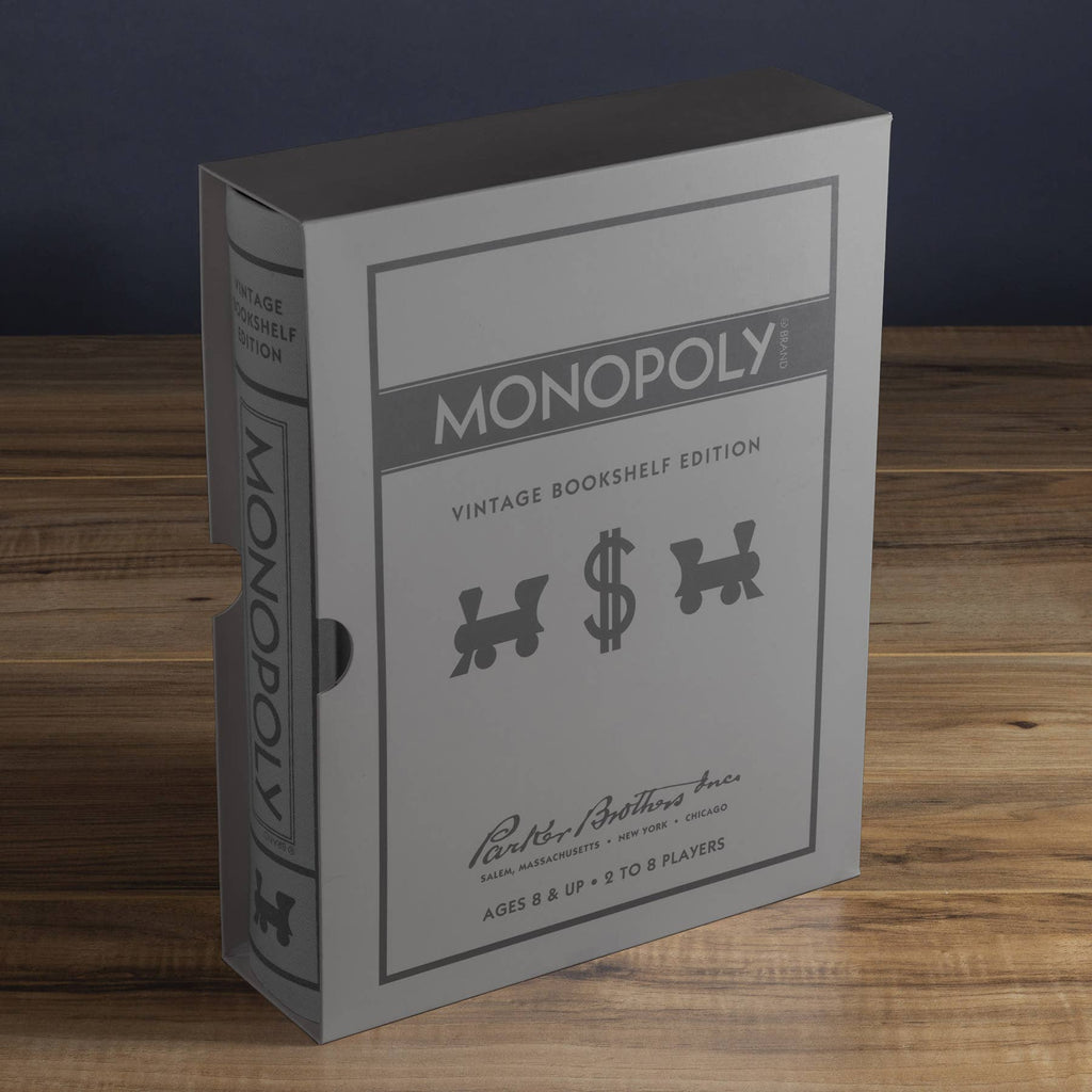 A Monopoly Vintage Bookshelf Edition box stands upright on a wooden surface, styled to look like a classic book in silver and gray with black graphics and premium components inside.