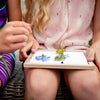 Two children’s hands and legs are visible as they hold the Huckleberry Pressed Flower Frame, displaying pressed blue and yellow blooms on white paper, resting on one child’s lap.