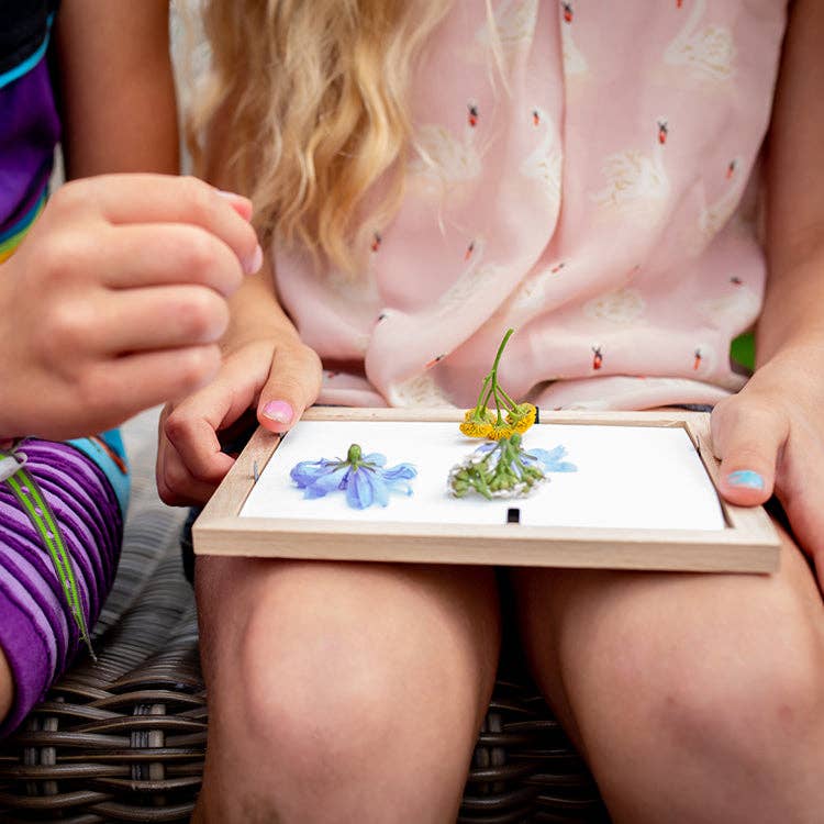 Two children’s hands and legs are visible as they hold the Huckleberry Pressed Flower Frame, displaying pressed blue and yellow blooms on white paper, resting on one child’s lap.