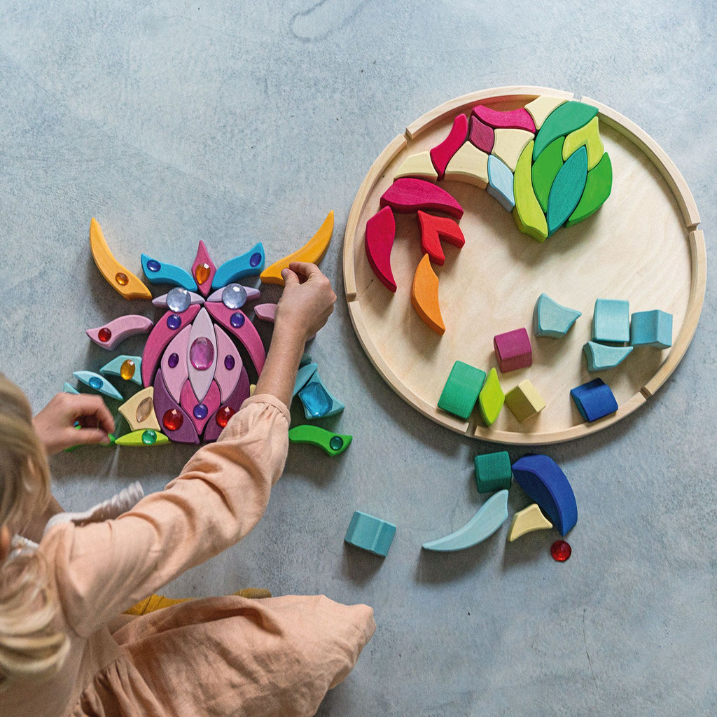 A child plays with colorful, abstract wooden puzzle pieces on the floor, enhancing their fine motor skills. The Grimm's Lara Circle Flower Building Set includes various shapes and vibrant colors, some assembled on a wooden base and others scattered around. The child wears a light-colored outfit and has blonde hair.