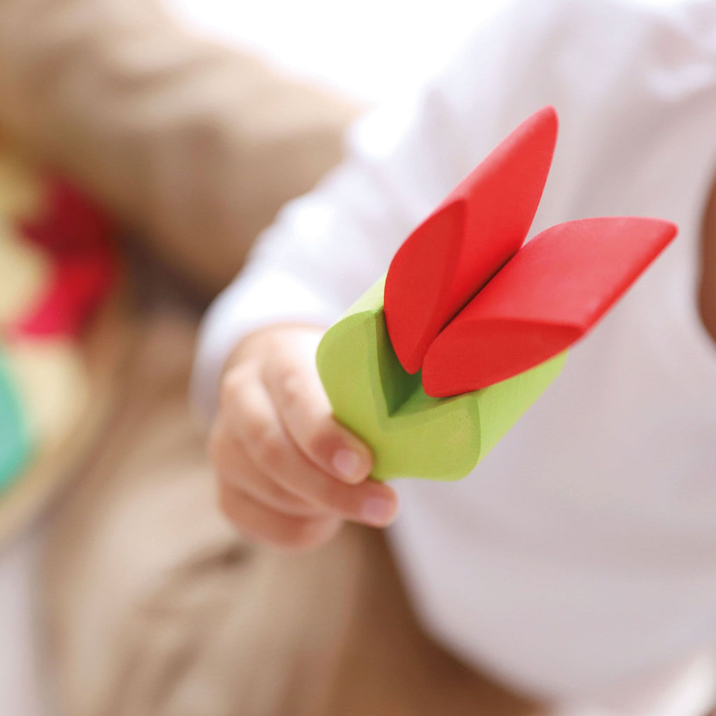 Close-up of a child holding a Grimm's Lara Circle Flower Building Set, with red petals and a green stem. The child is wearing a white shirt, and the background is blurred, focusing attention on the toy in the child's hand—a perfect way to enhance fine motor skills.