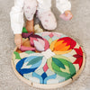 A child is playing with a colorful wooden puzzle on a gray carpet. The puzzle, resembling a Grimm's Lara Circle Flower Building Set, forms a circular pattern with petal-like pieces in red, blue, green, and yellow. As the child selectively places one of the pieces, they are honing their fine motor skills and spatial thinking skills.