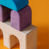 Grimm's Small Building Set - Floors features colorful wooden blocks in blue, purple, and natural tones with arch-shaped pieces on the bottom row, stacked into creative structures against a vibrant orange background.