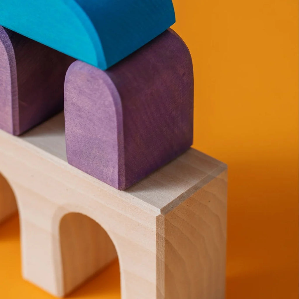 Grimm's Small Building Set - Floors features colorful wooden blocks in blue, purple, and natural tones with arch-shaped pieces on the bottom row, stacked into creative structures against a vibrant orange background.