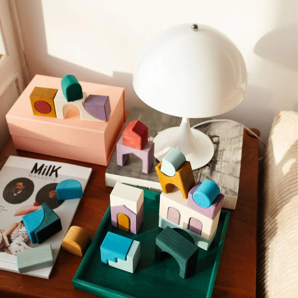 A wooden table displays Grimm's Small Building Set - Floors with colorful blocks, a modern white dome lamp, a pale pink box, a "Milk" magazine, and a green tray as sunlight streams through the window.
