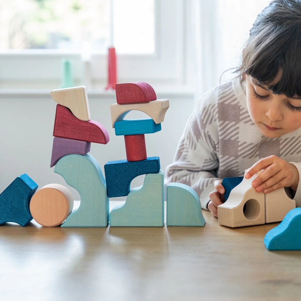 A young child focuses on stacking pieces from the Grimm's Small Building Block Set - Wave, building creative structures at a table as sunlight pours in through a window in the background.