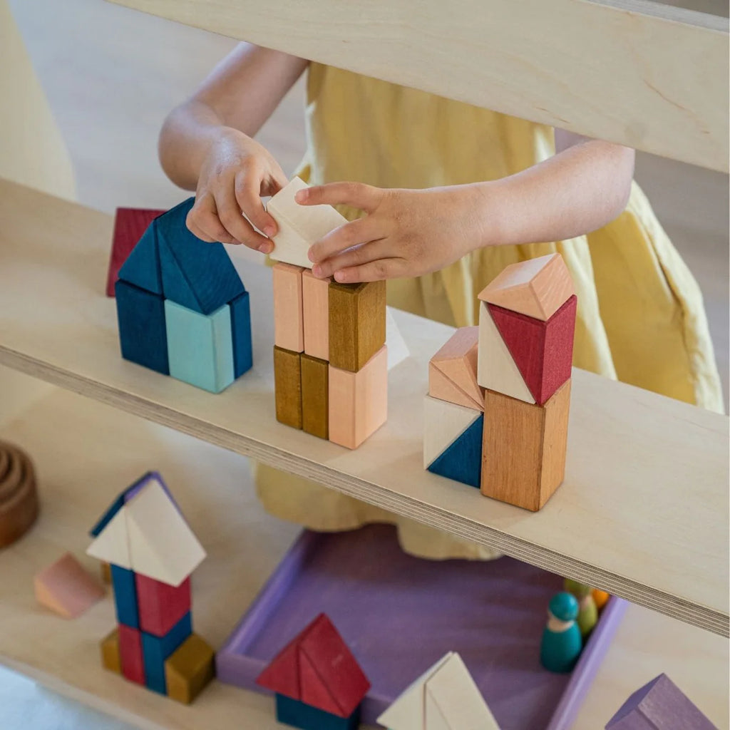 A child in a yellow dress builds with Grimm's Small Building Block Set - Rhythm on a shelf. Colorful towers and houses, resembling artistic sculptures, create a playful tiny art collection nearby.