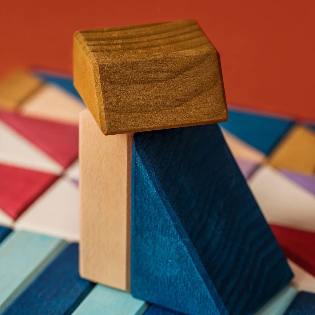A close-up of colorful wooden blocks from Grimm's Small Building Block Set - Rhythm, featuring a yellow block on top, a beige rectangular block in the center, and a blue triangular block at the bottom against a patterned background.