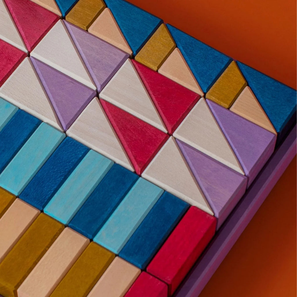 Grimm's Small Building Block Set - Rhythm features colorful wooden triangles and rectangles in blue, purple, gold, red, pink, and white arranged in a symmetric pattern on an orange background.