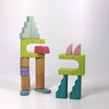 Two whimsical figures made from colorful solid wood blocks and shapes stand on a white background. One figure has a pastel blue and green body with pink details, while the other has a green body with a purple and pink head, resembling playful characters from an imaginative free-building adventure using the Grimm's Building World Cloud Play Block Set.