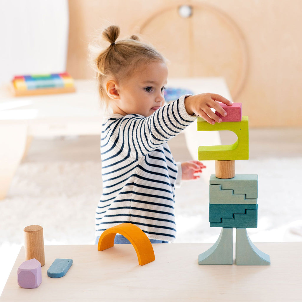 A toddler with blonde hair in pigtails, wearing a striped shirt, carefully stacks the Grimm's Building World Cloud Play Block Set at a light-colored table. In the background, there are more blocks and a white, minimalist interior—perfect for imaginative free building.