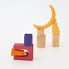 Grimm’s Building World - Desert Sand blocks arranged into two structures, including a small peg person inside a U-shaped piece and a yellow crescent moon balanced on pillars for imaginative free building.
