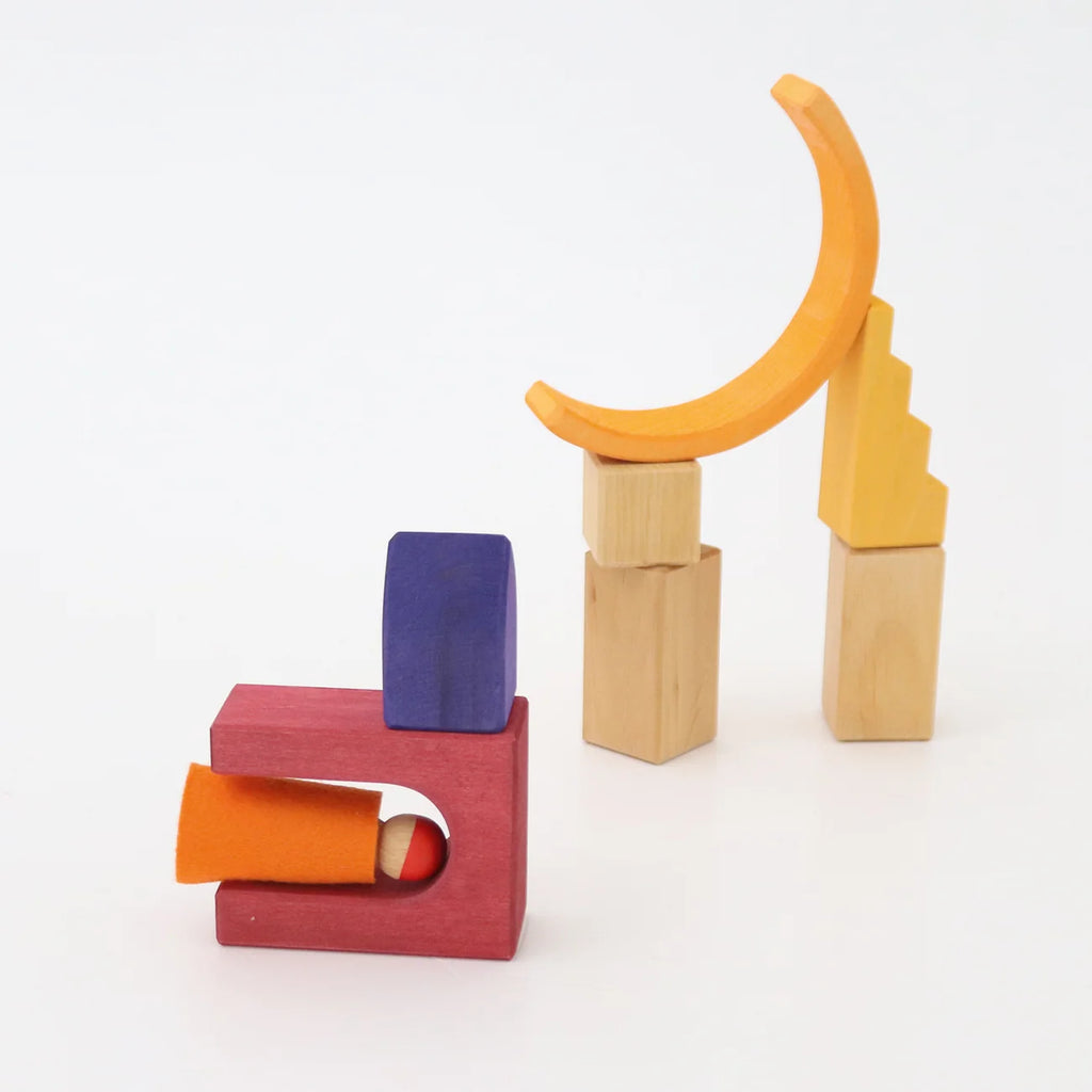 Grimm’s Building World - Desert Sand blocks arranged into two structures, including a small peg person inside a U-shaped piece and a yellow crescent moon balanced on pillars for imaginative free building.