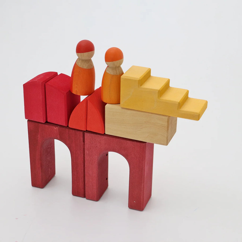 Two wooden peg people sit atop a structure made of red, orange, and yellow geometric Grimm’s Building World - Desert Sand wooden blocks.