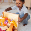 A child kneels on a rug, lifting a wooden tray of Grimm’s Building World - Desert Sand, featuring colorful geometric blocks for imaginative free building.