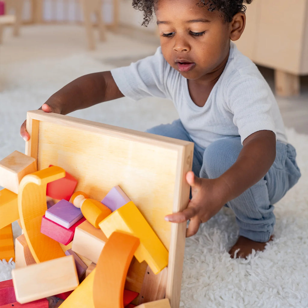 A child kneels on a rug, lifting a wooden tray of Grimm’s Building World - Desert Sand, featuring colorful geometric blocks for imaginative free building.