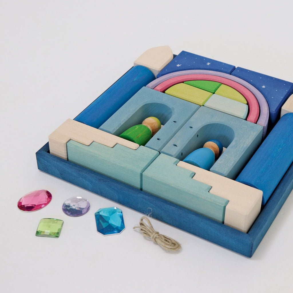 A colorful wooden toy set, arranged in a blue tray, features shapes resembling a castle with arches, towers, and a rainbow. Beside the tray are six shiny gem-like pieces in varying colors and shapes, along with a small length of string—perfect for creating small Grimm's Building World Polar Light Block Set.