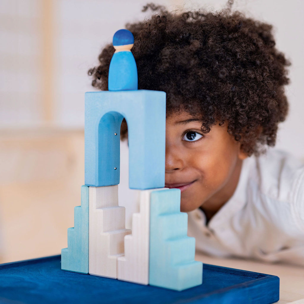 A young child with curly hair intently looks at a blue and white wooden block structure from their small Grimm's Building World Polar Light Block Set, topped with a small blue figure. The child appears focused and intrigued by their creation, which includes an arch and stairs-like design built from these imaginative building materials.