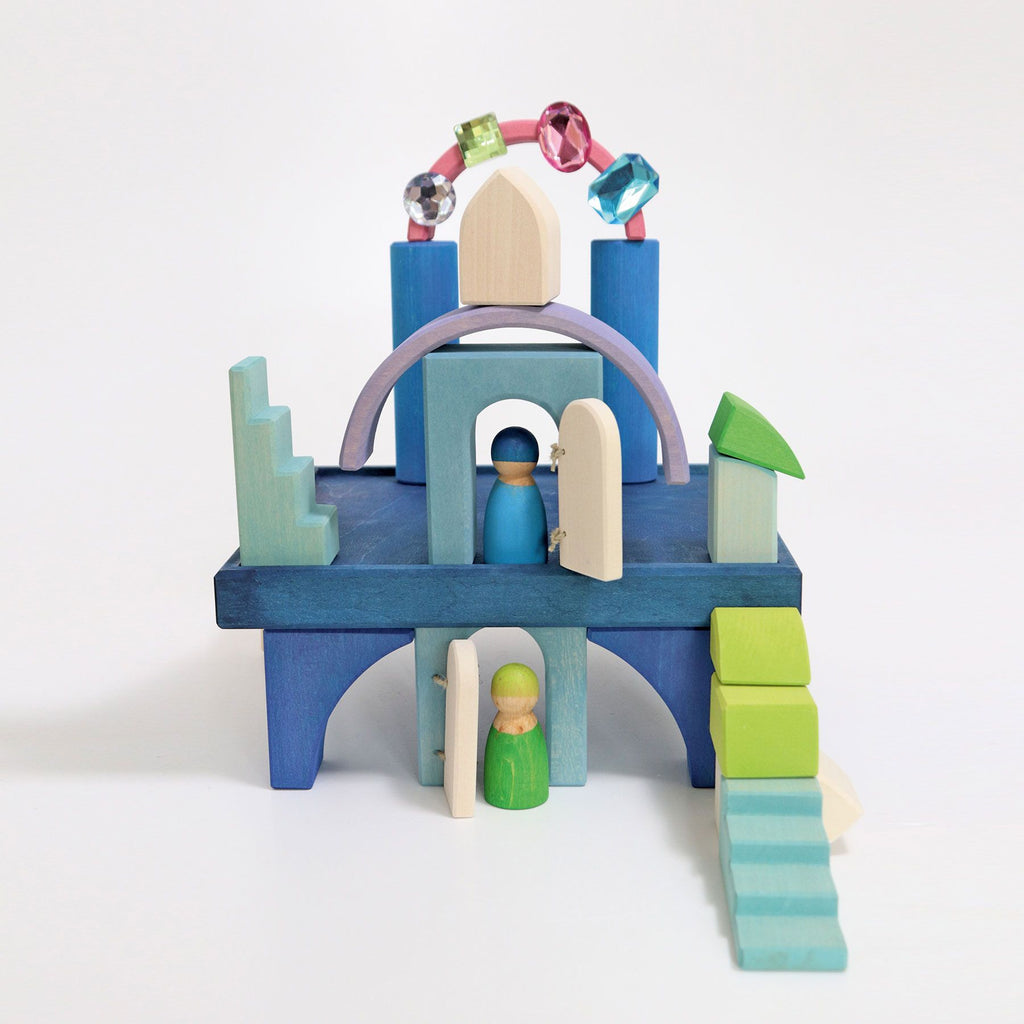 A colorful wooden castle playset, part of the Grimm's Building World Polar Light Block Set series, is arranged on a white background. It includes various blocks in blue, green, and purple with small doors which are open. Two wooden peg dolls, one in blue and one in green, are placed inside this delightful small Play World.