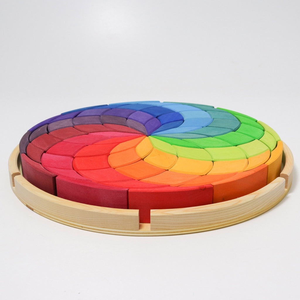 A Grimm's Wooden Color Spiral Block Set with colorful pieces arranged in a gradient pattern from red to purple. Perfect for young builders, the puzzle pieces fit into each other to form a compact circular shape, encased in a light wooden frame, enhancing fine motor skills and spatial thinking skills.