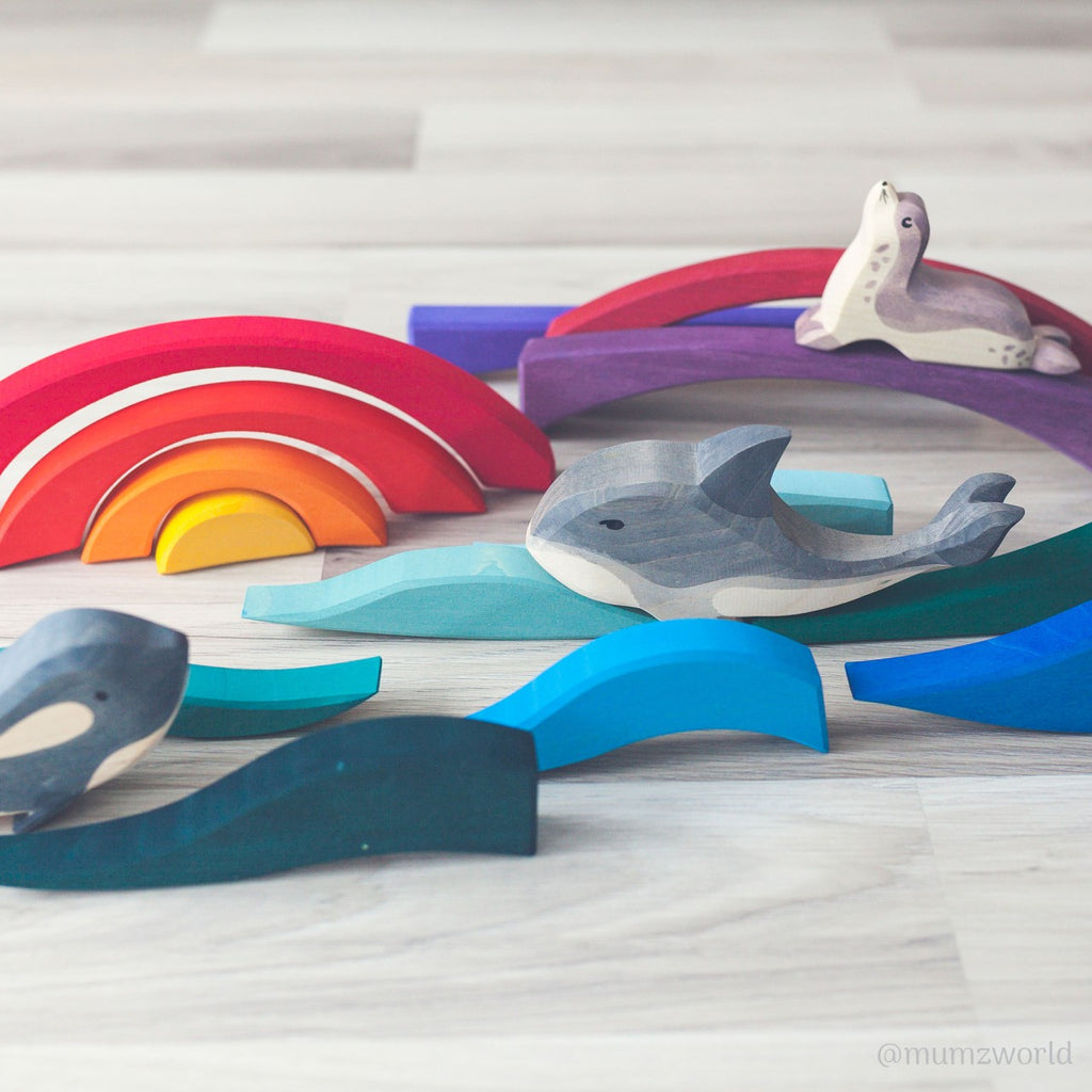 A playful arrangement of wooden toys is shown, designed to enhance fine motor skills. It includes colorful arches resembling a rainbow, wave-like pieces in various shades of blue, and animal figures of a seal and whale. The Grimm's Creative Puzzle Landscape Block Set is set on a light wooden floor. The image is bright and cheerful.