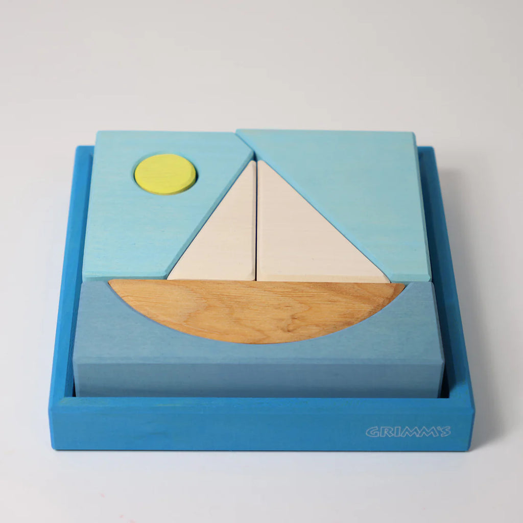 The Grimm's Building Set - Blue is a wooden puzzle featuring a sailboat with a yellow sun, blue sky, white sail, and light brown hull. Set in a blue tray labeled “GRIMM’S,” it’s perfect fun for toddlers and an ideal introduction to building blocks.