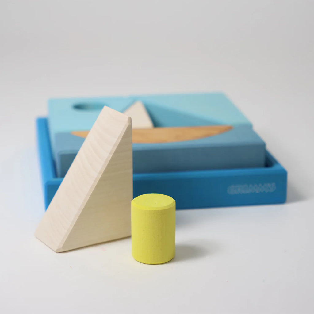 Grimm's Building Set - Blue features wooden blocks, including a yellow cylinder and a light triangular prism, arranged before a blue tray filled with blue and natural shapes—an ideal choice for any toddler toy collection.