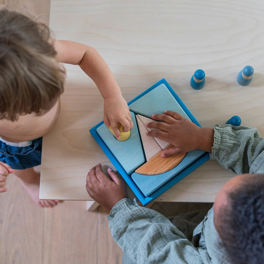 Two children sit at a wooden table playing with Grimm's Building Set - Blue. One child places a yellow piece into the colorful geometric puzzle while the other steadies it with both hands.