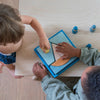 Two children sit at a wooden table playing with Grimm's Building Set - Blue. One child places a yellow piece into the colorful geometric puzzle while the other steadies it with both hands.