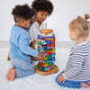 Three young children sit on a white carpet playing together with the Grimm's Assembly Set Musical Ball Run, watching marbles roll down its colorful spiral tracks while enjoying this engaging educational toy.