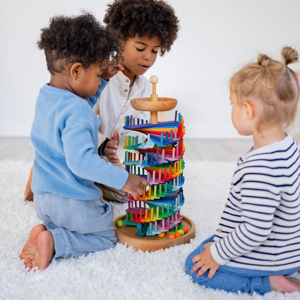 Three young children sit on a white carpet playing together with the Grimm's Assembly Set Musical Ball Run, watching marbles roll down its colorful spiral tracks while enjoying this engaging educational toy.