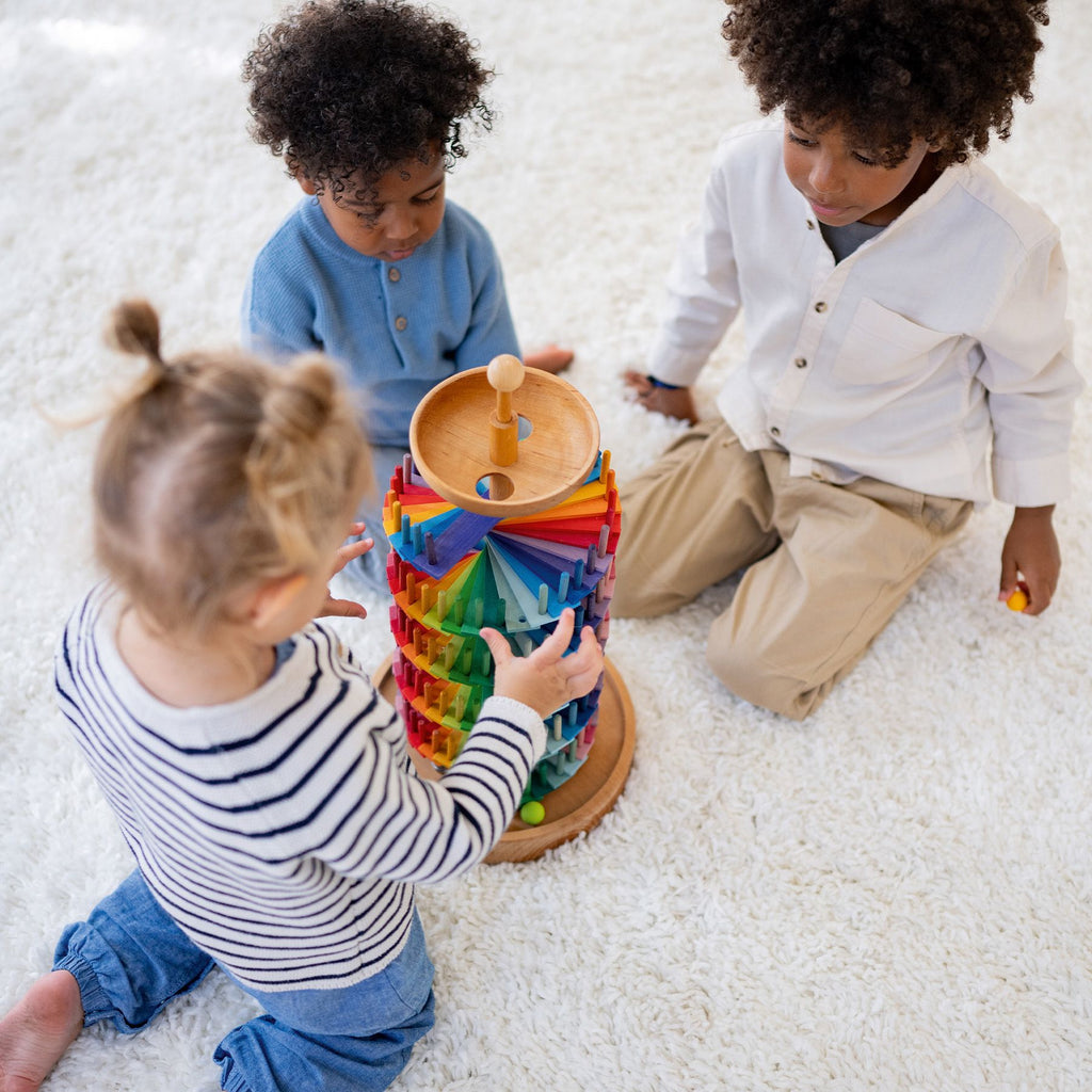 Three young children sit on a white carpet, playing together with the Grimm's Assembly Set Musical Ball Run, assembling the colorful wooden tracks and sending marbles racing down the tower.
