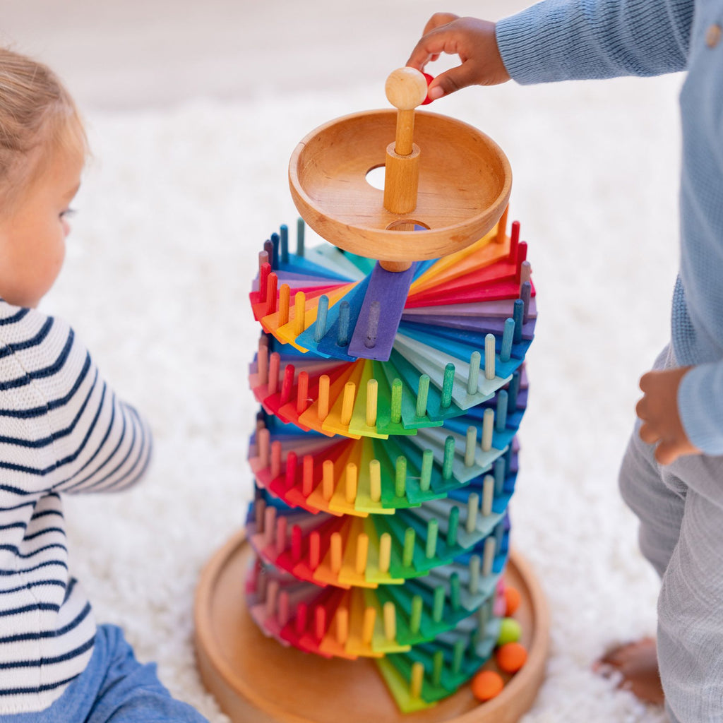 Two children play with Grimm's Assembly Set Musical Ball Run. One child drops a marble into the top spiral, watching as ramps and pegs create a vibrant rainbow pattern while the marble rolls down this educational wooden toy.