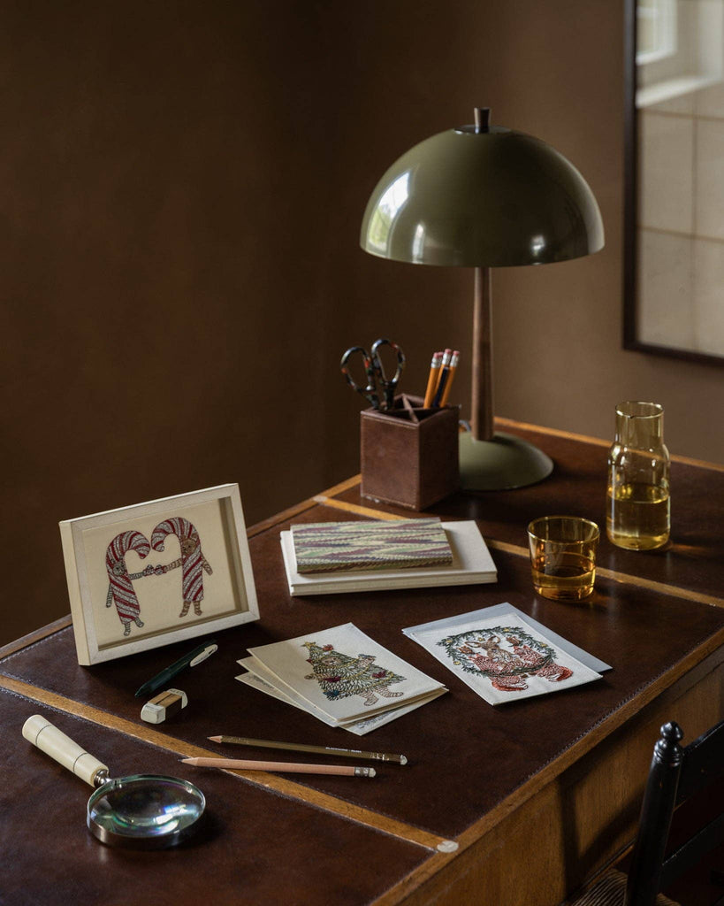 A wooden desk featuring a Coral & Tusk Christmas Tree Bear card illustration of two bears holding candy canes, with additional holiday-themed greeting cards, a lamp, and writing supplies nearby.