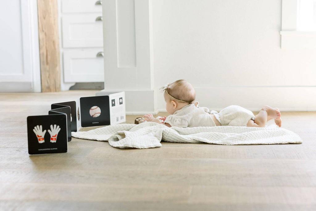 A baby enjoys tummy time on the floor, engaged with Baby's Tummy Time Sports—black and white sports-themed flashcards featuring images like gloves and a baseball—in a bright, minimal room for visual stimulation.