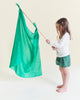 A young child with long hair stands barefoot in a white long-sleeve shirt and green shorts, holding Sarah's Silks Solid Color Playsilk - Emerald on a wooden stick. The plain, light-colored background highlights this open-ended play accessory.