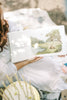 A person in a white dress sits on a blanket outdoors, holding open Winnie-the-Pooh: Finding Feelings—a board book showing a bear and child under a tree, perfect for toddlers to explore emotions.