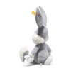 The Steiff Looney Tunes Bugs Bunny is a soft gray and white plush rabbit sitting upright, with large ears, white gloves, one arm extended, and a yellow Steiff Button in Ear tag on its left ear.