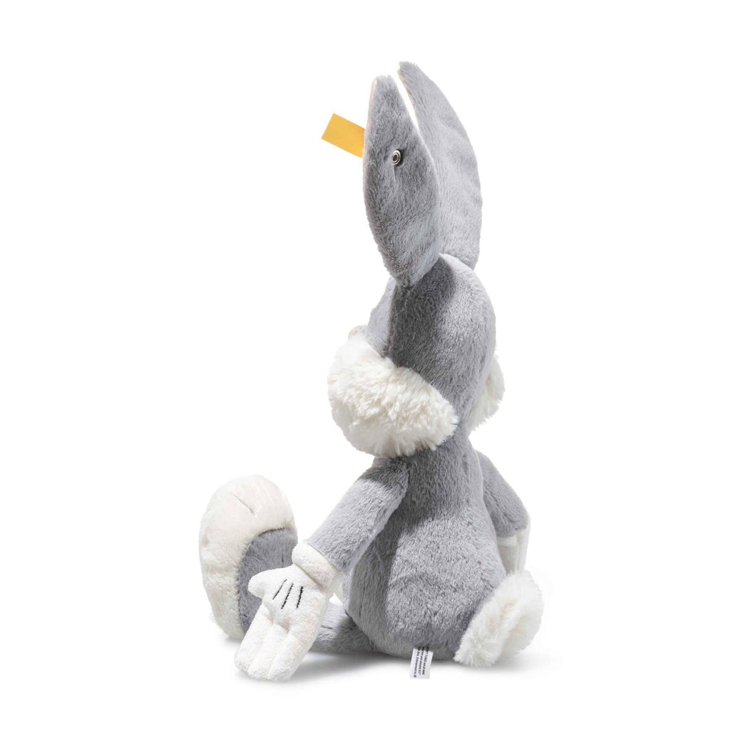 The Steiff Looney Tunes Bugs Bunny is a soft gray and white plush rabbit sitting upright, with large ears, white gloves, one arm extended, and a yellow Steiff Button in Ear tag on its left ear.