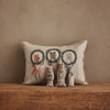 The Christmas Wreath Trio Pocket Pillow, a beige cushion with embroidered fox, bear, and rabbit inside festive wreaths, sits on a wooden surface. In front are three plush woodland friends that match the pillow’s animal designs.