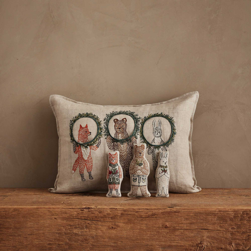 The Christmas Wreath Trio Pocket Pillow, a beige cushion with embroidered fox, bear, and rabbit inside festive wreaths, sits on a wooden surface. In front are three plush woodland friends that match the pillow’s animal designs.
