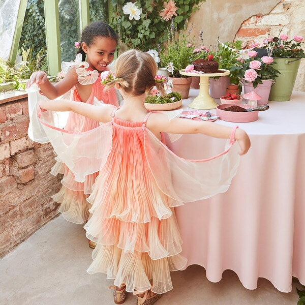 Two young girls wearing the Meri Meri Fairy Costume - Final Sale, featuring layered, pastel-colored fairy dresses, are playfully posing with outstretched arms in front of a decorated table. The table is adorned with cakes, pastries, and flowers, creating a whimsical, fairy-like atmosphere in the sunlit room.